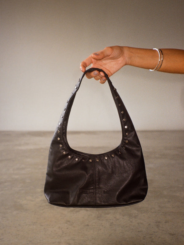 Vista frontal mini bolso negro de cuero sintético con taches metálicos. 