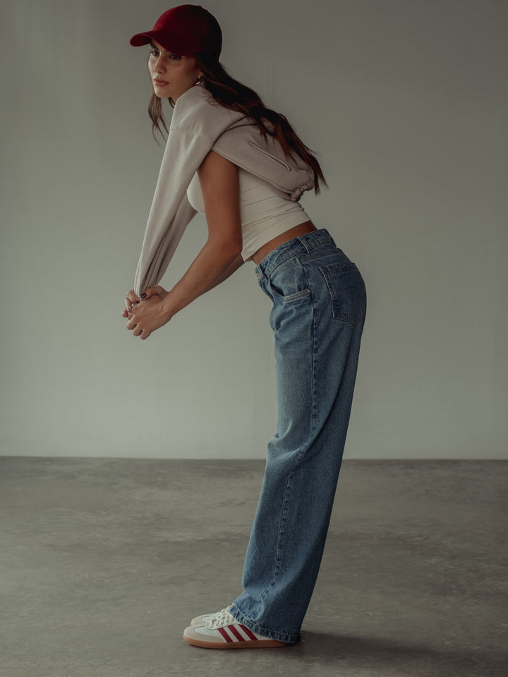 Vista lateral de la modelo usando un jean, camiseta blanca, gorra y buzo en su espalda color arena