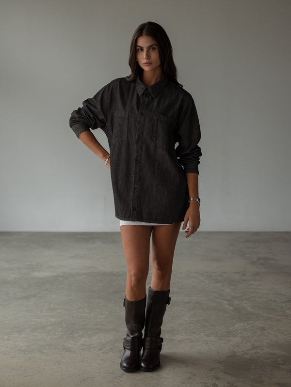 Modelo usando camisa negra oversized con botones cerrados, mini falda y botas. 