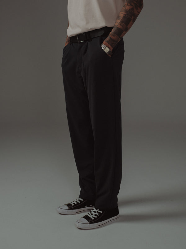 Vista lateral modelo con pantalón negro, cinturón de cuero y tenis.