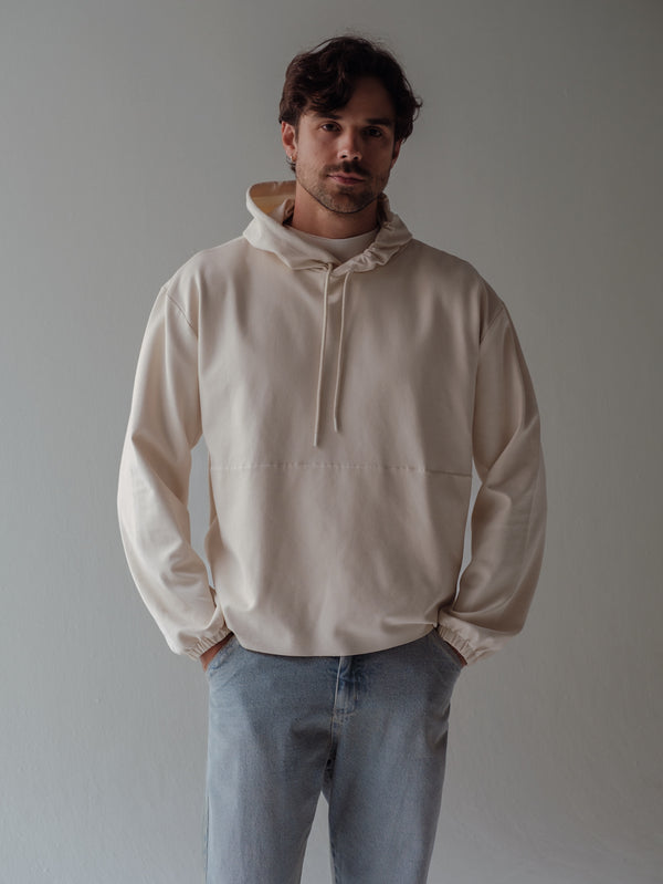 Modelo con manos en los bolsillos usando buzo crema con capucha. 