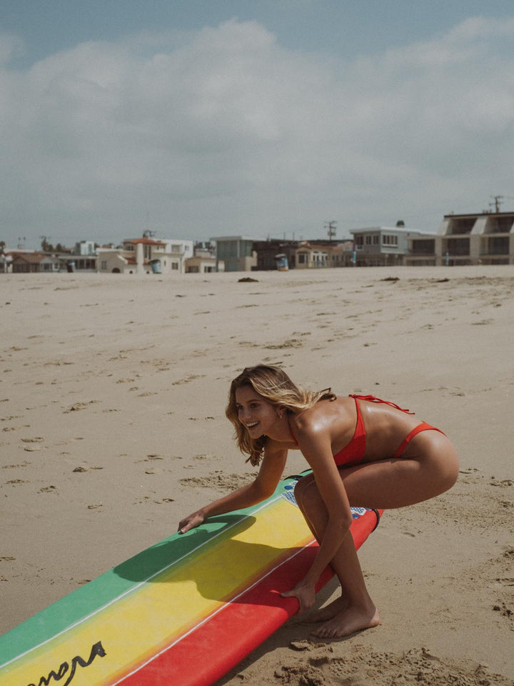 La modelo lleva set de vestido de baño en color rojo mientras tiene en su mano una tabla de surf