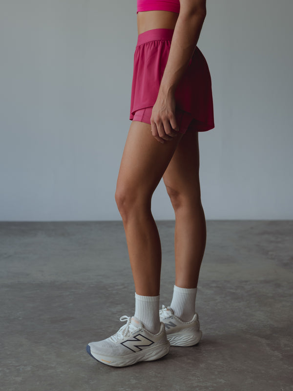 Modelo parada de perfil usando short deportivo fucsia, medias blancas y tenis.