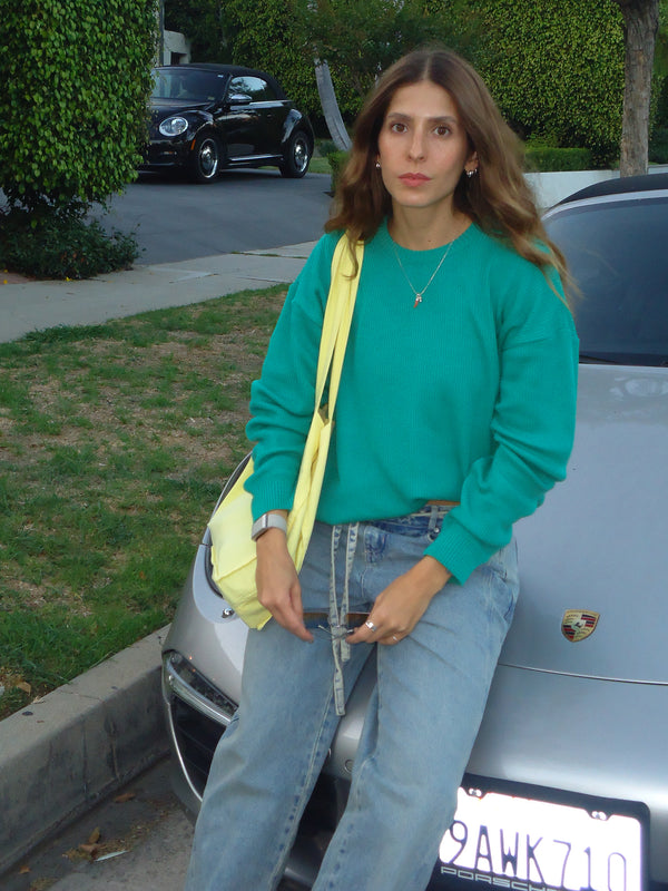 Modelo recostada en un carro usando suéter tejido verde y jean.