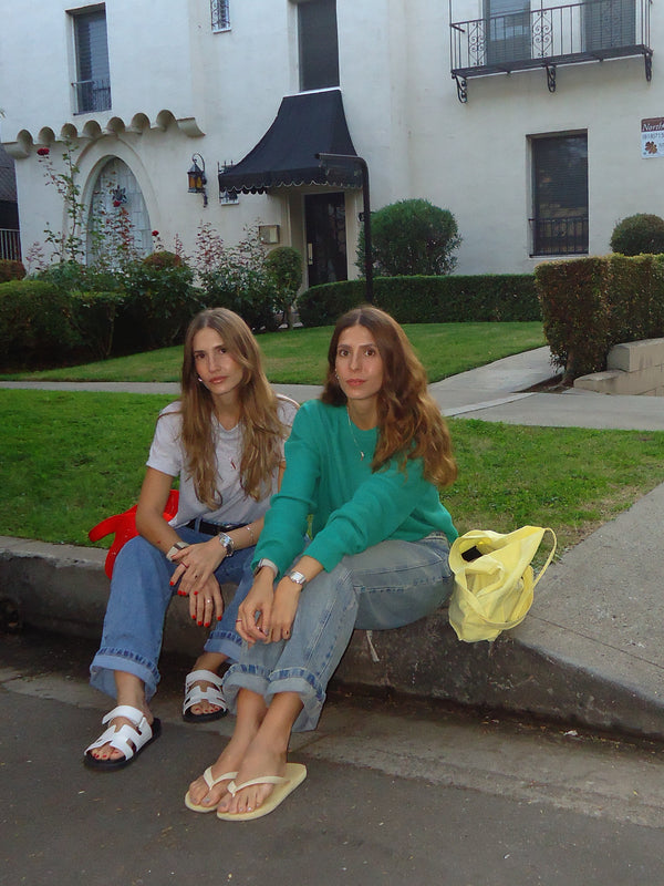 Dos modelos sentadas en la calle usando ropa casual con sandalias.