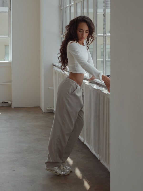 Modelo de pie recostada a ventana vistiendo pantalón gris y top manga larga crudo.