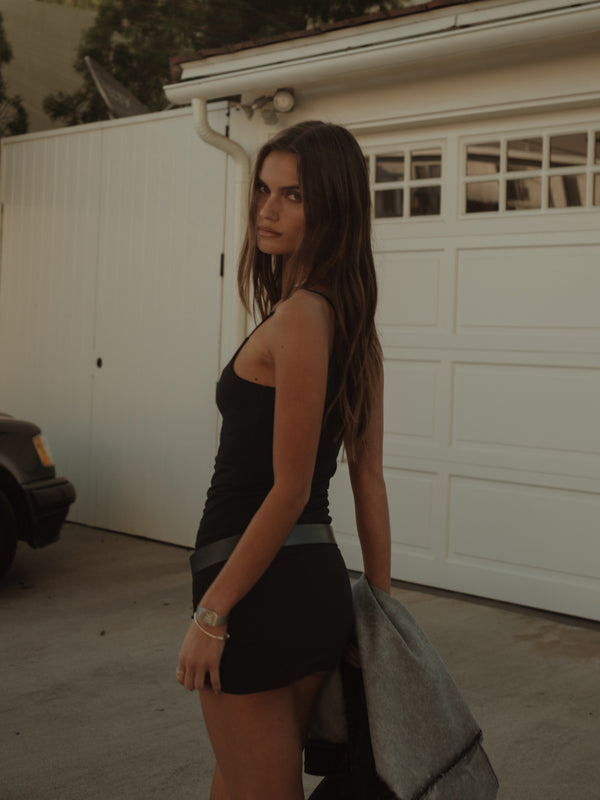 Modelo de lado usando mini vestido negro sosteniendo una chaqueta. 