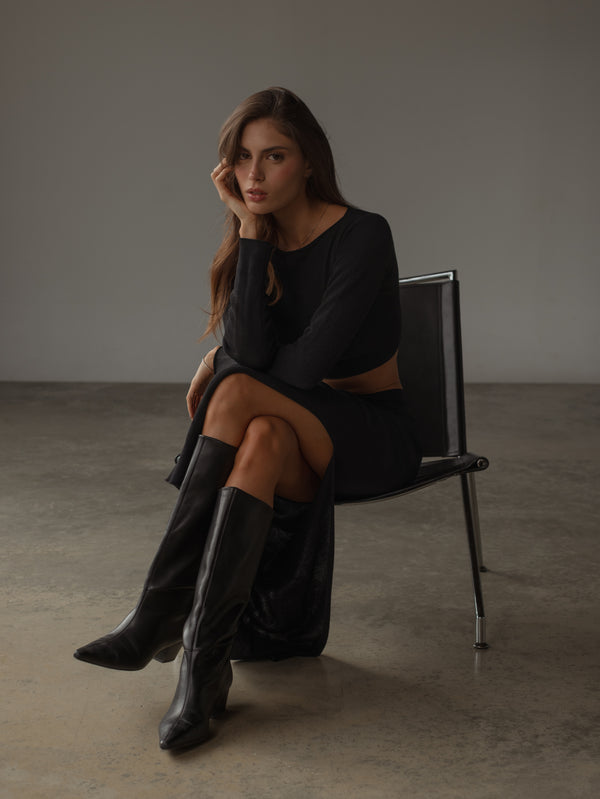 Modelo sentada usando vestido negro tejido y botas negras.